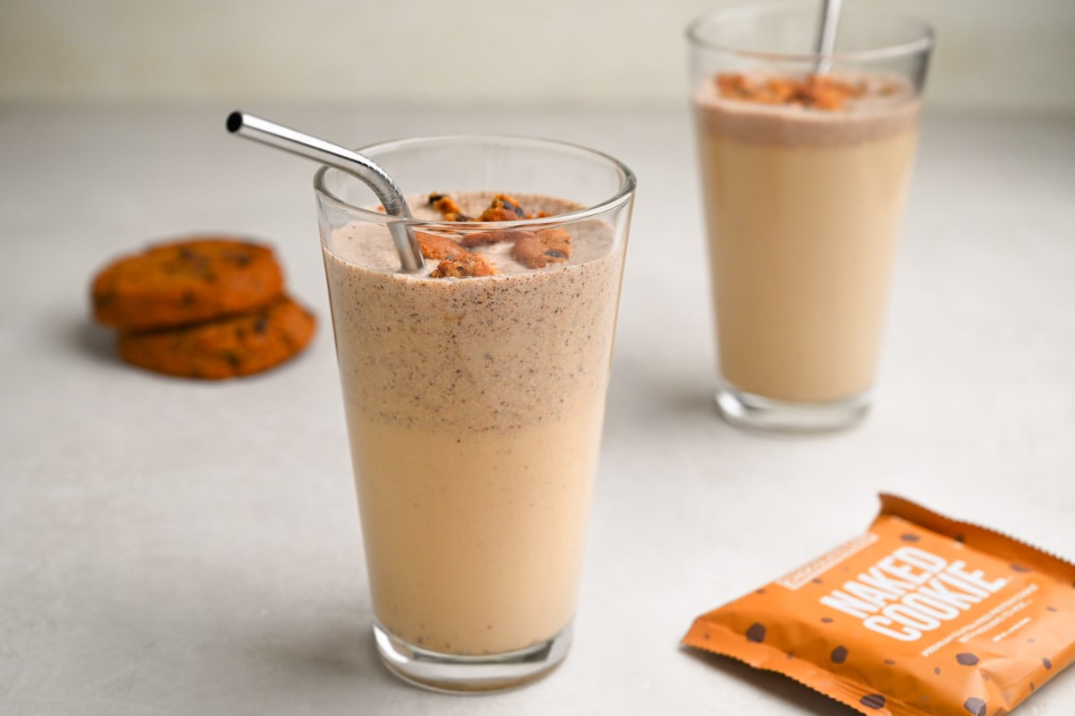 Two glasses of cookies and cream protein shakes with cookie pieces on top, metal straws and cookies in the background.