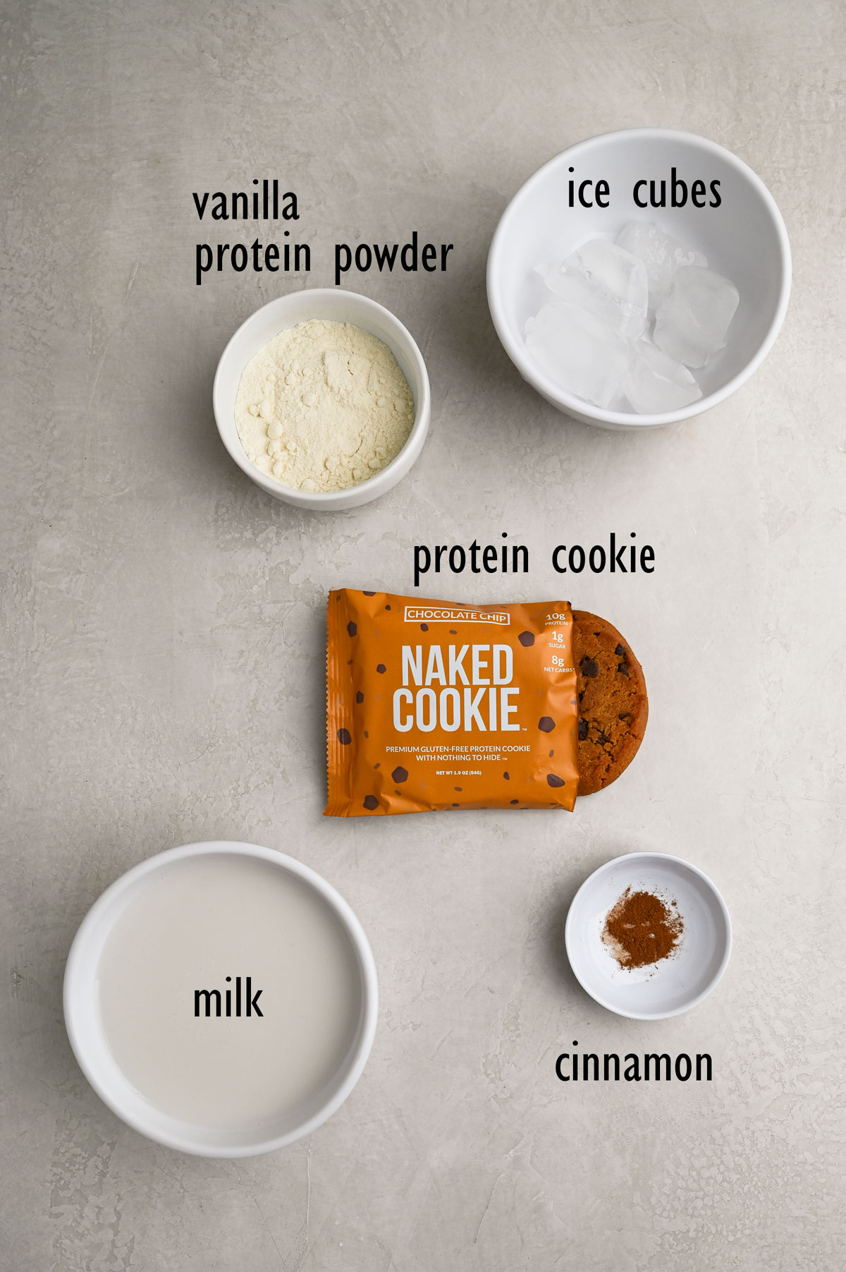 Shake ingredients including protein powder, a protein chocolate chip cookie, ice cubes, almond milk and cinnamon.