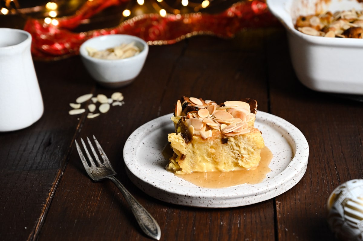 Straight on view on a plate of panettone bread pudding topped with almonds and brandy sauce on dark wood with the pan and Christmas lights in the background.