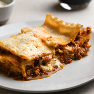 Close up, straight on view of a slice of lasagna bolognese on a plate with wine and parm in the background.