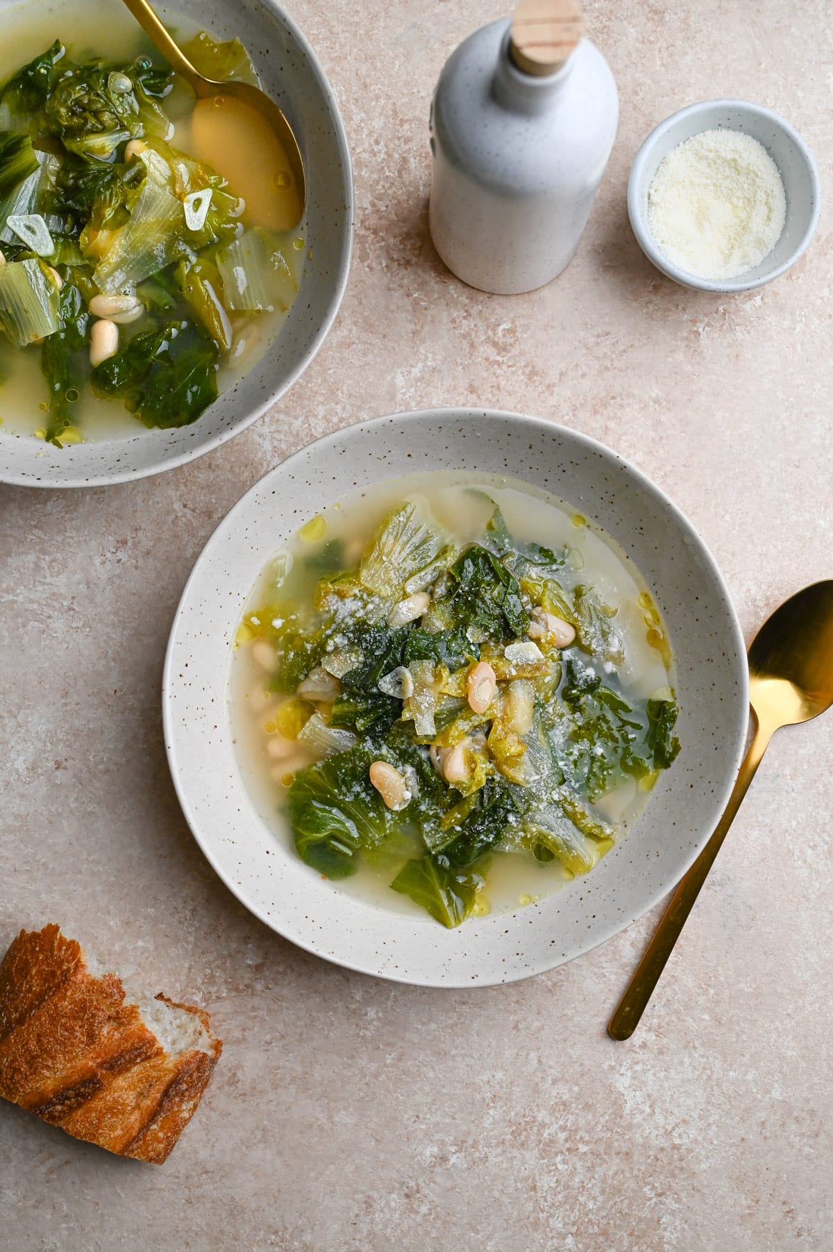 Overhead view of two bowls of Escarole and Beans surrounded by parmesan, a spoon and a piece of bread.