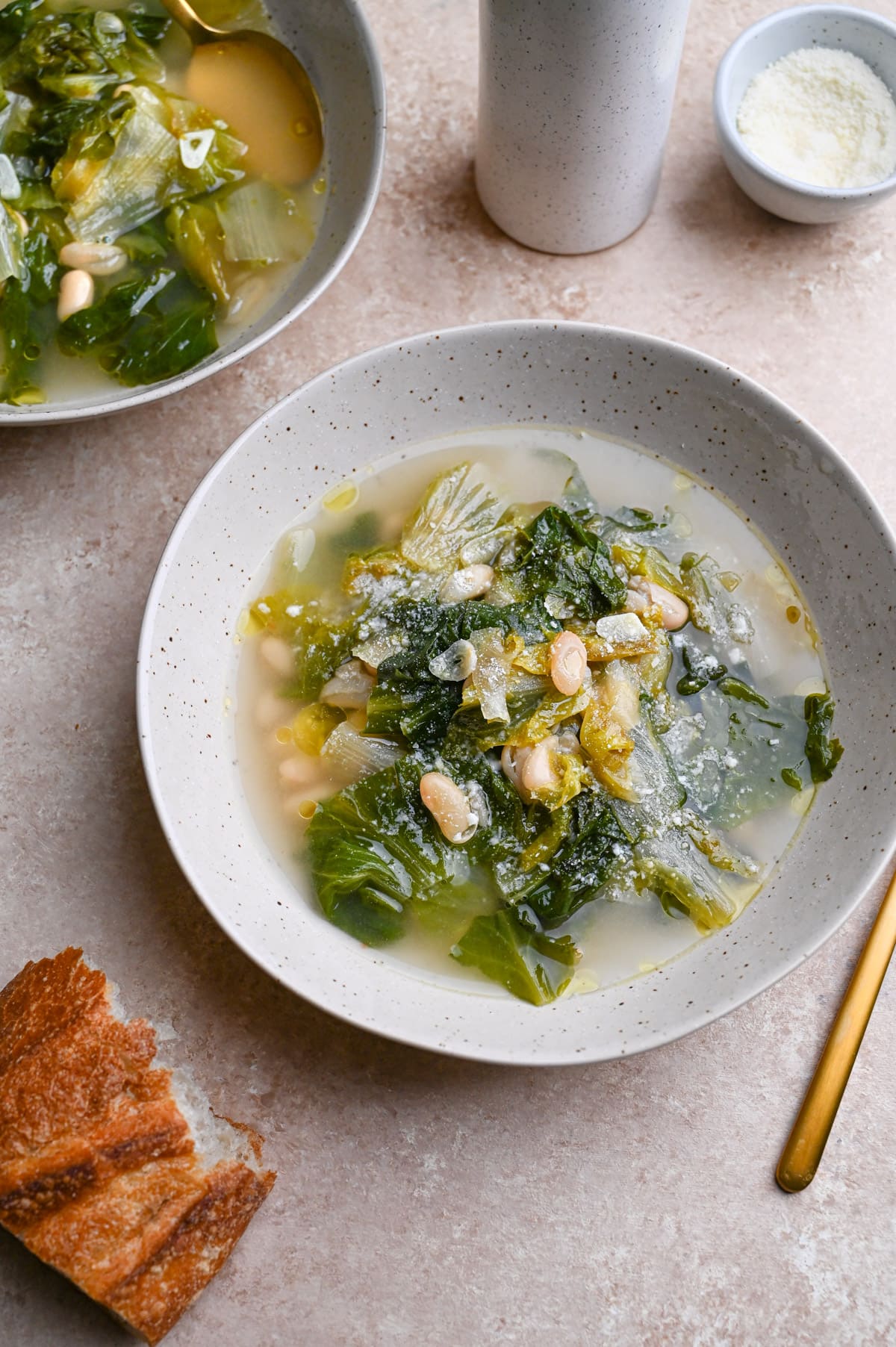Angled view of two bowls of Escarole and Beans surrounded by parmesan, a spoon and a piece of bread.
