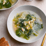 Angled view of two bowls of Escarole and Beans surrounded by parmesan, a spoon and a piece of bread.