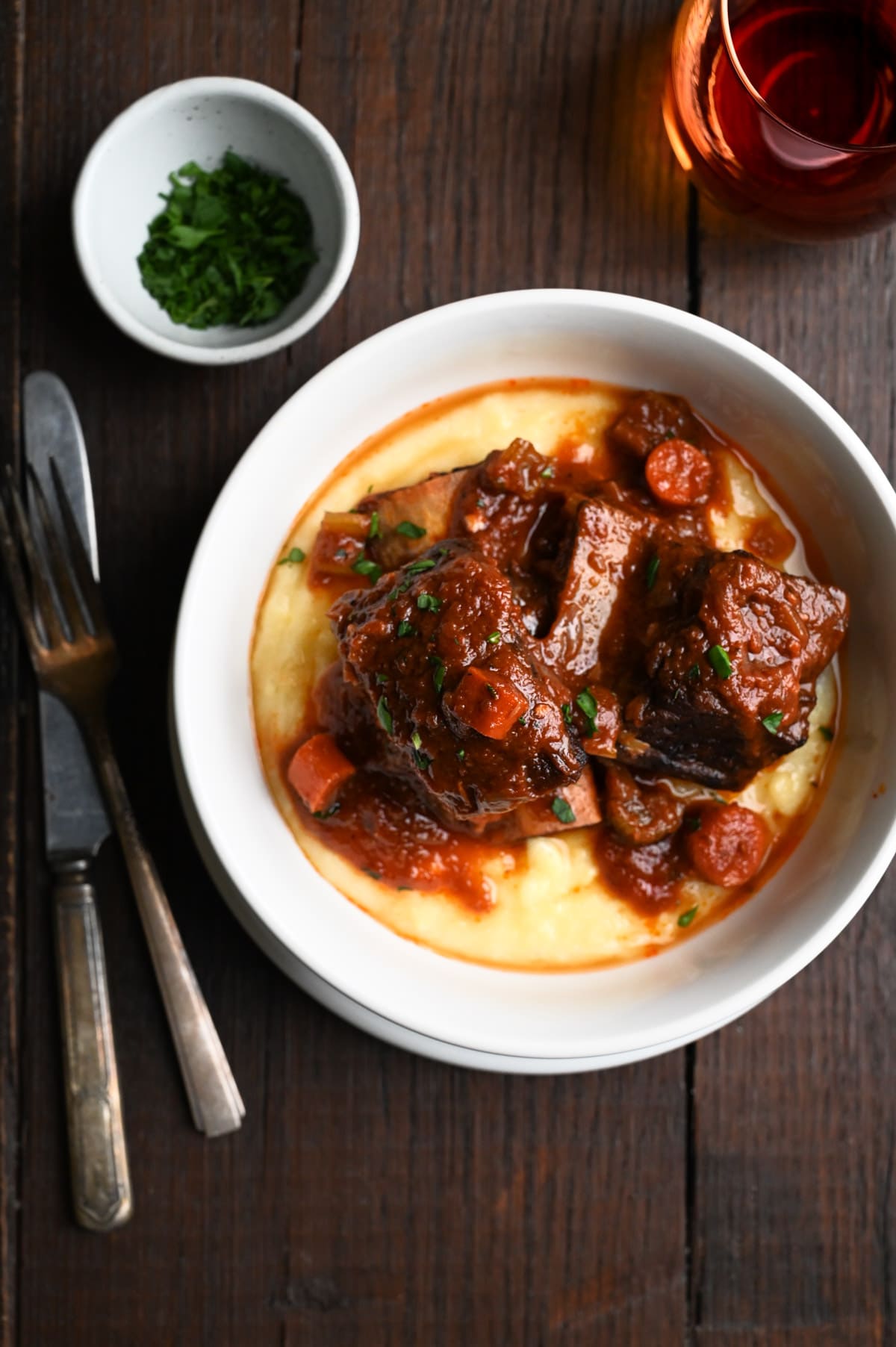 Overhead view of a bowl of polenta topped with Italian Short Ribs on a dark wood surface.