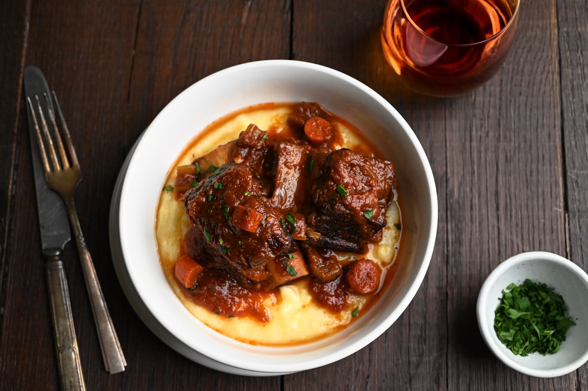Overhead view of a bowl of polenta topped with Italian Short Ribs on a dark wood surface.