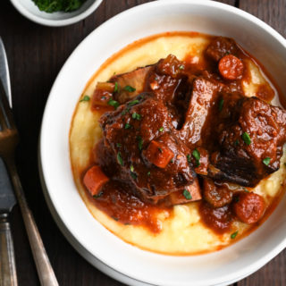 Overhead view of a bowl of polenta topped with Italian Braised Short Ribs on a dark wood surface.