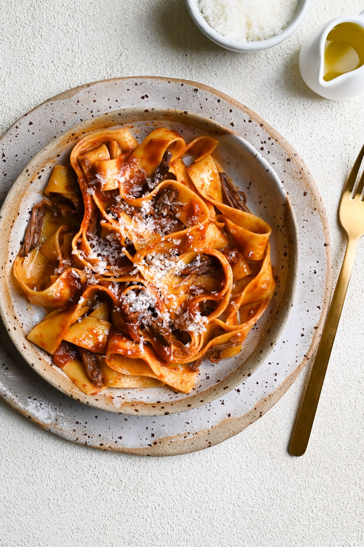 Short Rib Ragu with Pappardelle Pasta topped with parmesan on a textured, cream surface.