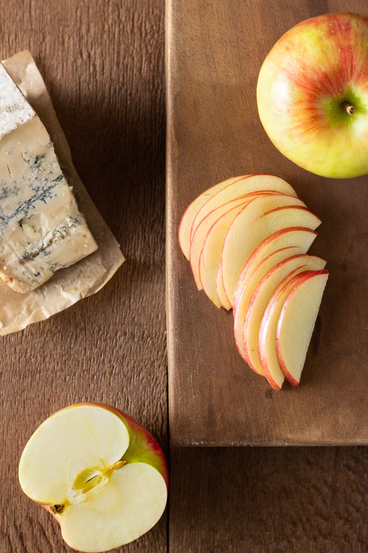 Overhead view of whole and sliced apples and a wedge of blue cheese on a cutting board on a wood surface.