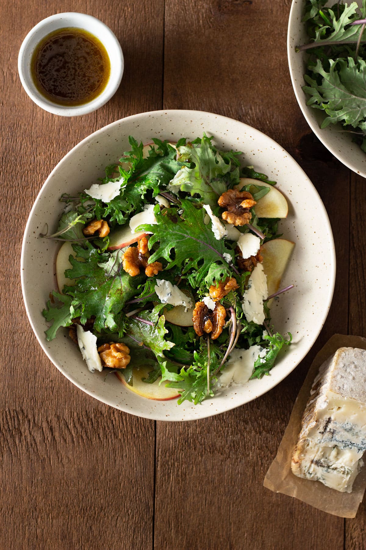 Overhead view of Kale and Apple Salad with Walnuts and Blue Cheese next to a bowl of balsamic dressing on a wood surface.