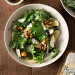 Overhead view of Kale and Apple Salad with Walnuts and Blue Cheese next to a bowl of balsamic dressing on a wood surface.