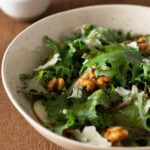 Close up, angled view of Kale and Apple Salad with Walnuts and Blue Cheese on a wood surface.