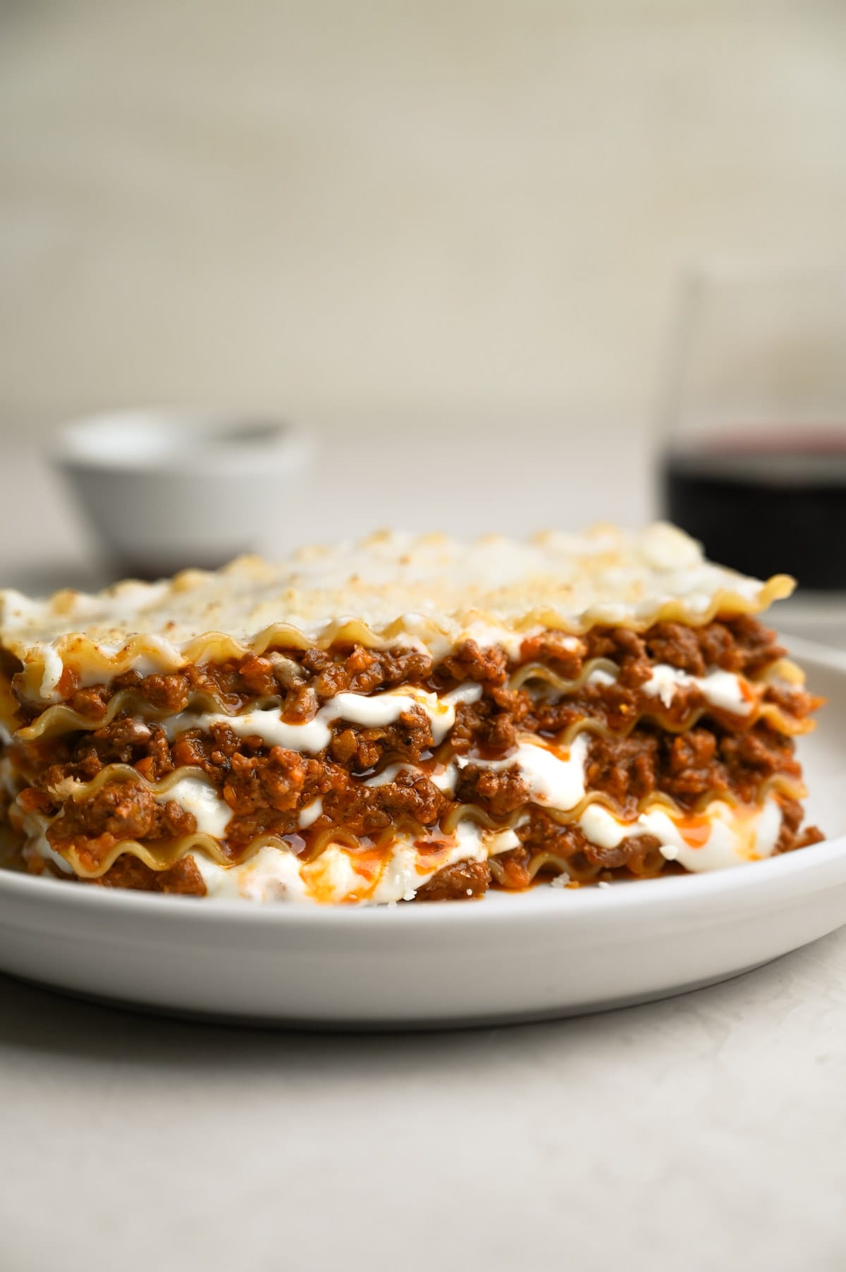 Close up of a plate of Lasagna Bolognese on a cream surface surrounded by a glass of wine, a fork and a bowl of parmesan.