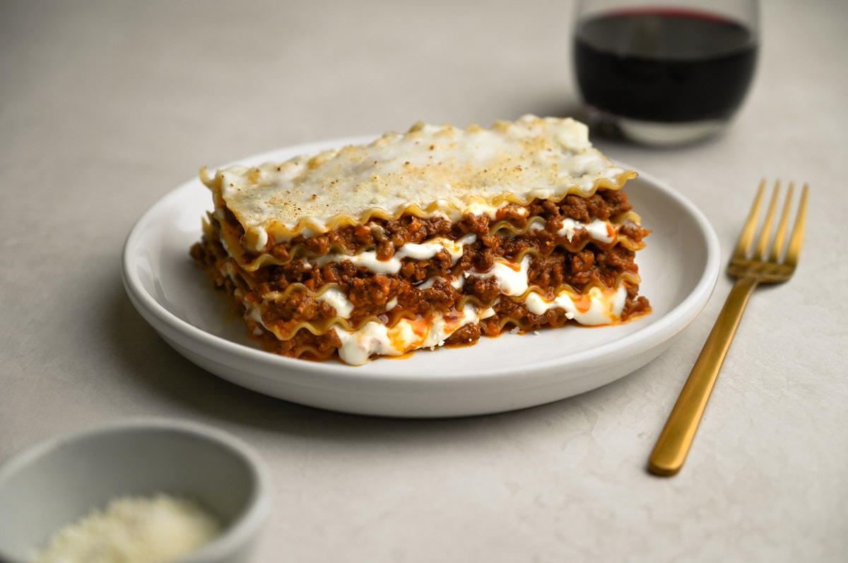 A plate of Lasagna Bolognese with Bechamel Sauce on a cream surface surrounded by a glass of wine, a fork and a bowl of parmesan.