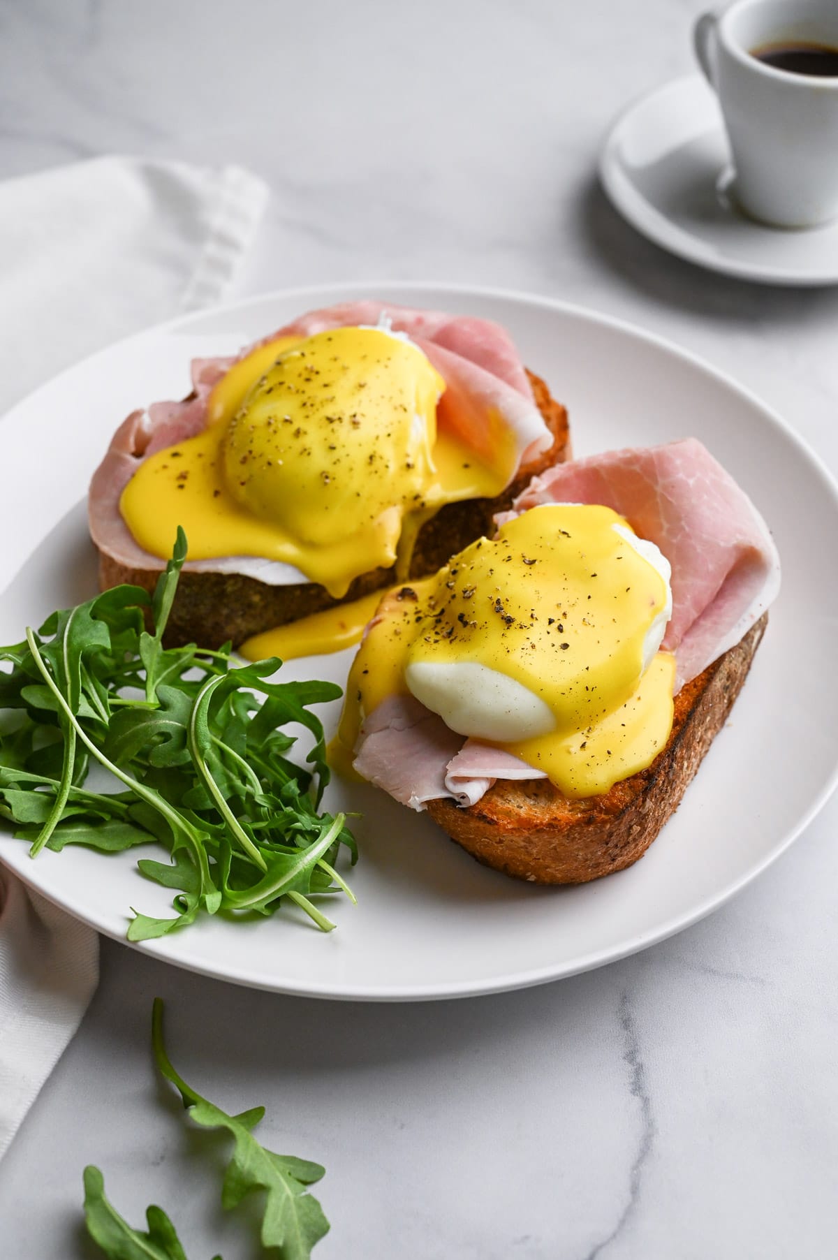 Angled view of toast topped with Italian ham, poached eggs and hollandaise sauce with arugula on the side on a marble surface.