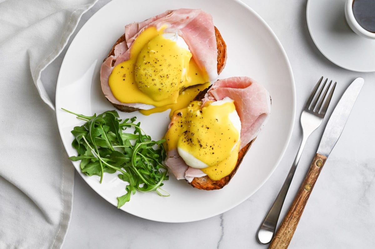 Overhead view of Italian Eggs Benedict with arugula on a marble surface next to a cup of espresso and a fork and knife.
