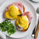 Overhead view of Prosciutto Eggs Benedict with arugula on a marble surface next to a cup of espresso and a fork and knife.