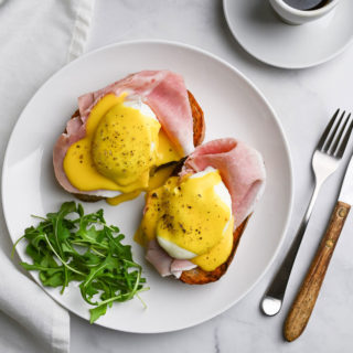 Overhead view of Italian Prosciutto Eggs Benedict with arugula on a marble surface next to a cup of espresso and a fork and knife.