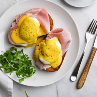 Overhead view of Italian Prosciutto Eggs Benedict with arugula on a marble surface next to a cup of espresso and a fork and knife.